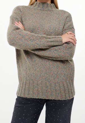 Personne portant un pull en maille gris moucheté, bras croisés, associé à un pantalon noir à petits pois blancs.