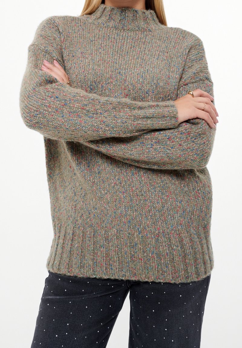 Personne portant un pull en maille gris moucheté, bras croisés, associé à un pantalon noir à petits pois blancs.