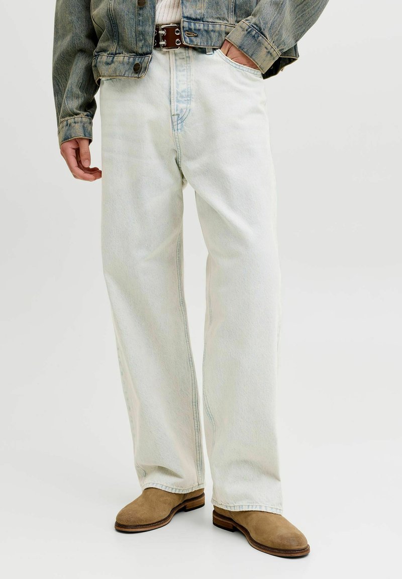 Persona che indossa jeans a gamba larga azzurri, stivali in camoscio marroni, giacca di jeans e cintura marrone con la mano sinistra in tasca.