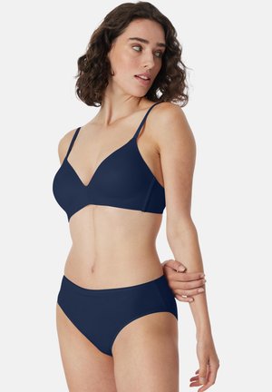 Vrouw met krullend bruin haar, gekleed in een bijpassende marineblauwe bh en ondergoedset, kijkt naar links met één arm vastgehouden door de andere.