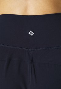 Dunkelblaue Sportleggings mit einer glatten Textur, Flatlock-Nähten und einem weißen Emblemsdesign am Bund für feinere Details.