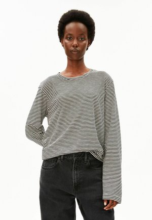 Persoon met kort krullend haar, gekleed in een zwart-wit gestreept shirt met lange mouwen en een zwarte spijkerbroek, staand tegen een witte achtergrond.