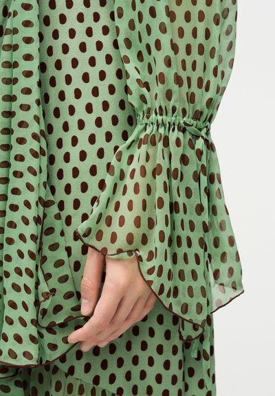 Χέρι ακουμπισμένο σε πράσινο διάφανο ύφασμα με καφέ πουα, που διαθέτει μανίκι με βολάν, φουσκωτό και με ελαστική μανσέτα.