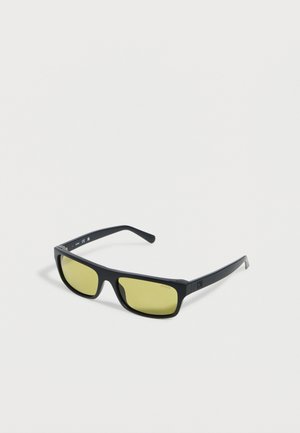 Sorte rektangulære solbriller med guletonede linser, vinklet på en hvid baggrund.