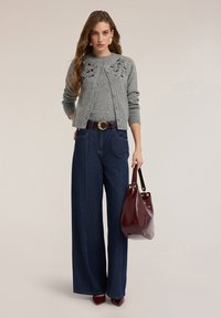 Šedý kardigan s korálkovými detaily, doplněný o šedý top a tmavě modré džíny s širokými nohavicemi. Bordó kabelka, lodičky s špičatou špičkou a akcent zlatého pásku.