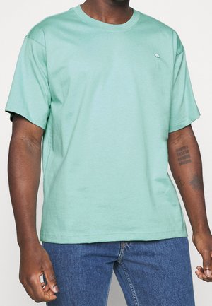 Mand iført en ensfarvet lys grøn kortærmet t-shirt og blå jeans med synlige tatoveringer på sin højre underarm.