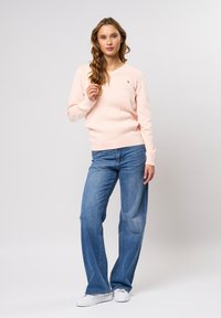 Světlě růžový žebrovaný svetr s výstřihem do V, kombinovaný se širokými modrými džínami a bílými teniskami, zdobený malým logem.