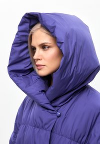 Jachetă pufoasă purpurie cu un guler supradimensionat și cu glugă, având o textură netedă și lucioasă. Dispune de o închidere cu butoane și un design matlasat.