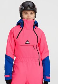 Felroze skikleding met blauwe accenten, voorzak met rits, verstelbare capuchon en elastische manchetten. Geprofileerd waterdicht materiaal.