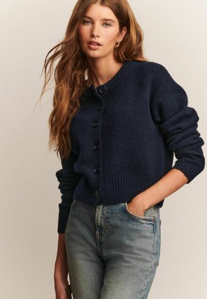 Frau mit langen, welligen Haaren, die eine marineblaue, geknöpfte Strickjacke und hellblaue, hoch taillierte Jeans trägt, mit einer Hand in der Tasche.