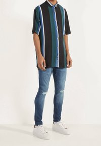 Camicia a maniche corte con bottoni, a righe verticali nere, turchesi e blu. Indossata con jeans blu strappati e sneakers bianche.