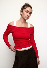 Roter, gerippter Schulterfreier Pullover mit langen Ärmeln, kombiniert mit einem weißen Hemd darunter, getragen zu hoch taillierter dunkler Hose.