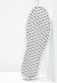 Semelle d’une basket blanche avec des motifs de bande de roulement en forme de diamant et d’hexagone, positionnée verticalement contre un mur d’angle blanc.