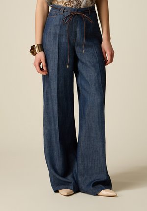 Donkerblauwe denim broek met wijde pijpen en plooien aan de voorkant, een dunne bruine riem in de taille gestrikt, gecombineerd met beige puntige schoenen en een gouden armband.