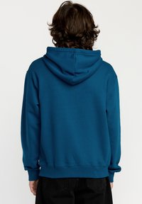 Sudadera azul verdosa con capucha ajustable, mangas largas, y puños y dobladillo acanalados. La vista desde atrás muestra un corte relajado y una textura suave.