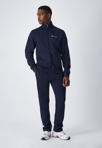 Chándal Navy fabricado en tejido suave, que incluye una chaqueta con cremallera y pantalones a juego con puños acanalados. Presenta un pequeño logo en el pecho.