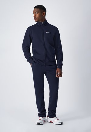 Chándal Navy fabricado en tejido suave, que incluye una chaqueta con cremallera y pantalones a juego con puños acanalados. Presenta un pequeño logo en el pecho.