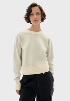 Young person with short wavy hair wearing a cream-colored knit sweater and black pants, standing against a plain light background.