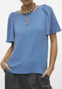 Blusa azzurro chiaro con texture, colletto a chiave e maniche a sbuffo corte, abbinata a una collana di perle argentate, indossata con pantaloni grigi.