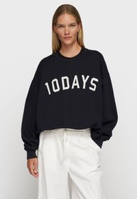 Schwarz oversized Sweatshirt mit weißem "10DAYS"-Logo, gerippter Rundhalsausschnitt und lockeren Ärmeln, kombiniert mit weißen Jogginghosen mit Kordelzug.