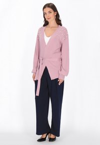 Ljusrosa cardigan med ribbat mönster, v-ringning, dekorerade axlar och midjeband, tillsammans med marinblå vida byxor och svarta klackar.