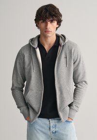 Grijze zip-up hoodie met een zachte textuur, voorzakken en een capuchon met trekkoord, gedragen over een zwart poloshirt, in combinatie met lichtdenim jeans.
