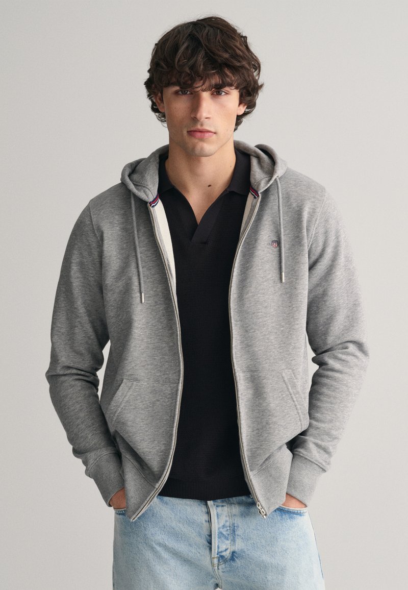 Grijze zip-up hoodie met een zachte textuur, voorzakken en een capuchon met trekkoord, gedragen over een zwart poloshirt, in combinatie met lichtdenim jeans.
