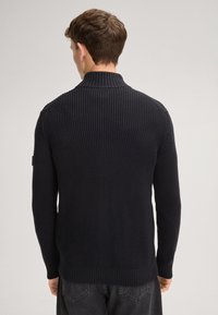 Zwarte gebreide trui met een hoge kraag, geribbelde manchetten en zoom; gestructureerd patroon zichtbaar op de achterkant.