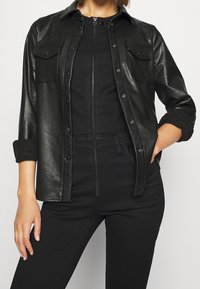 Czarna kurtka koszulowa z ekologicznej skóry z dwiema kieszeniami na klapie, zapięciem na guziki, podwiniętymi rękawami i gładką teksturą. Nosi się ją na czarnym kombinezonie.