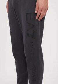Tmavé šedé jogger nohavice vyrobené z mäkkého materiálu, s veľkým čiernym nápisom "EA7" a "EMPORIO ARMANI" pozdĺž ľavej nohy.