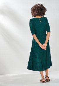 Grünes Kleid mit schwarzen Punkten, knielang, mit einem gestuften Design, Puffärmeln und einem rückseitigen Schlüssellochverschluss. Ein Paar flache Sandalen.