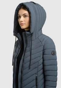 Sivá prešívaná bunda typu puffer s veľkou kapucňou, zipsom na prednej strane a dvoma zipsovými vreckami. Obsahuje čierne šnúrky a logo na rukáve.