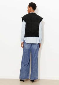 Haut noir sans manches en maille avec une finition texturée, porté sur une chemise boutonnée bleu clair, associé à un jean en denim bleu à jambes larges.