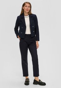 Marineblaues doppelt geknöpftes Blazer mit Struktur und silbernen Knöpfen, getragen über einem weißen Oberteil und kombiniert mit navyblauen Hosen und schwarzen Slippern.