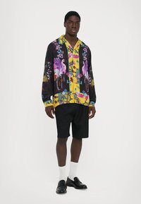 Cămașă cu un model floral colorat, având un design galben și negru, asortată cu pantaloni scurți negri. Modelul poartă șosete albe și pantofi negri.
