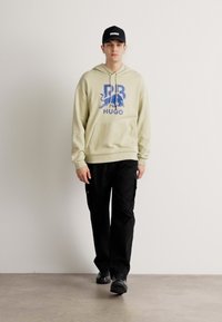 Felpa beige con grafica blu 'HUGO' e design di leone; abbinata a pantaloni cargo neri e sneakers nere; cappellino semplice con logo 'HUGO'.