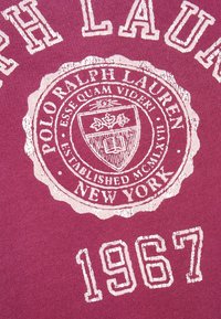 Ralph Lauren-logotyp med vapensköld, texten "POLO RALPH LAUREN NEW YORK", latinsk motto och året "1967" på blekt mörkrött tyg.