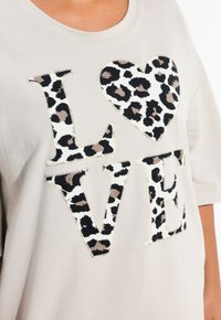 Hellbeige, übergroßes T-Shirt mit dem Aufdruck "LOVE" in fetten Buchstaben, ergänzt durch ein Herzdesign aus schwarzem und tanfarbendem Leopardenmuster. Aus weicher Baumwolle.