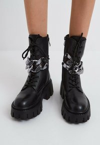 Bottes en cuir noir à la cheville avec une semelle épaisse, ornées d'une chaîne métallique argentée autour du haut et d'une fermeture à lacets.