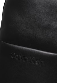 Zaino in pelle nera con una texture liscia e un leggero riflesso. Logo in rilievo ben visibile sulla parte anteriore con una finitura opaca a contrasto.