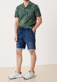 Hombre vestido con una camisa de manga corta con un estampado de hojas verdes, pantalones cortos de mezclilla azul con cordón y zapatillas blancas, de pie contra un fondo liso.