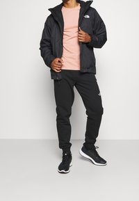 Černá nepromokavá bunda, růžové tričko, černé tepláky a černé tenisky. Hladké textury s akcenty loga na oblečení. Sportovní styl.