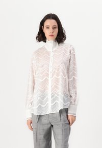 White lace blouse with a high collar, intricate wave pattern, button front, and long sleeves, paired with light gray checkered trousers.