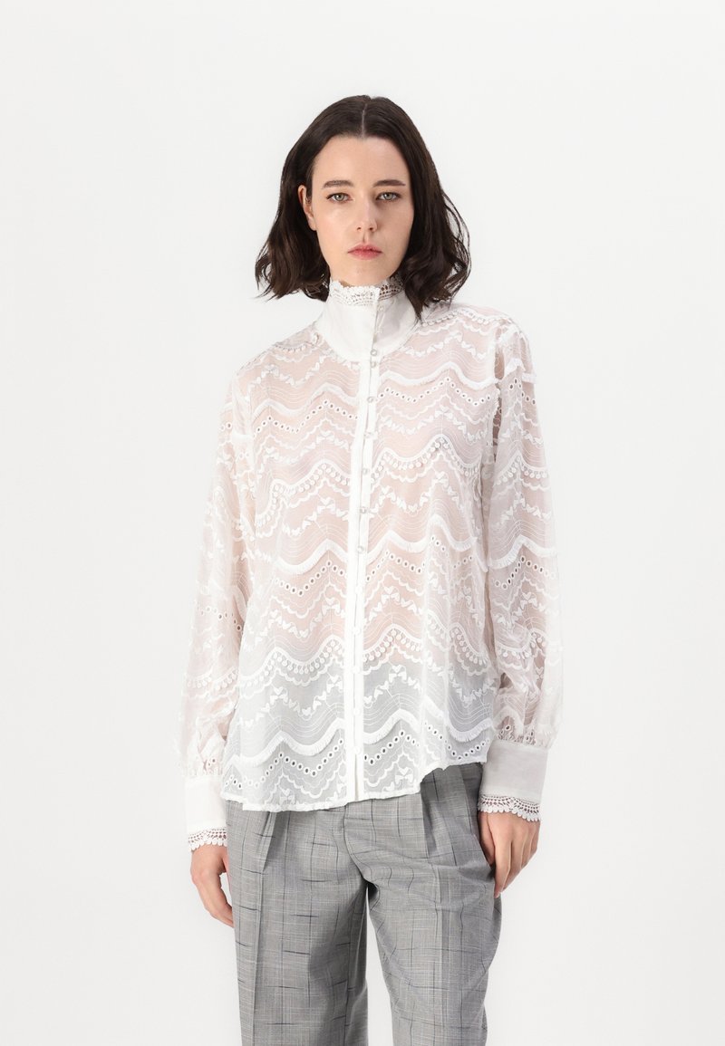 White lace blouse with a high collar, intricate wave pattern, button front, and long sleeves, paired with light gray checkered trousers.