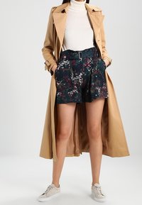 Manteau trench beige avec une taille ceinturée, porté par-dessus un short court vert foncé à motifs. Le mannequin porte des baskets claires et un col roulé blanc ajusté.