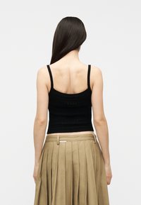 Γυναίκα με μακριά σκούρα μαλλιά φοράει μαύρο πλεκτό τοπ με λεπτές τιράντες και μπεζ πλισέ φούστα με ψηλή μέση, με την πλάτη γυρισμένη.