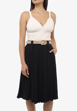 Donna che indossa un top in maglia bianco senza maniche, gonna midi nera plissettata e cintura beige, in piedi con una mano nella tasca della gonna.