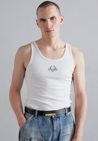 Top de tirantes blanco con textura de canalé que presenta un pequeño diseño gráfico negro en el pecho, combinado con pantalones cortos de mezclilla de tiro alto y un cinturón negro.