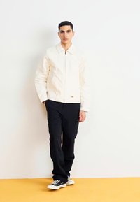 Cream jacket with a collared neckline, black cargo pants, and black sneakers, against a light background with an orange floor.