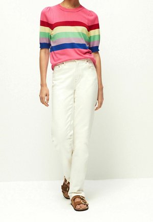 Femme portant un haut à manches courtes rayé coloré, un pantalon blanc taille haute et des sandales marron à boucle, debout devant un fond uni.